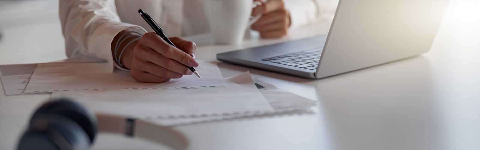 Person holding a pen, writing on papers beside a laptop, with a cup in the other hand.