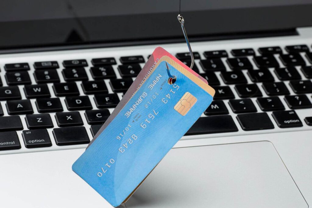 A credit card with the name "NAME SURNAME" and number "5439 1590 8430 0110" is hooked on a fishhook above the keyboard of a laptop.