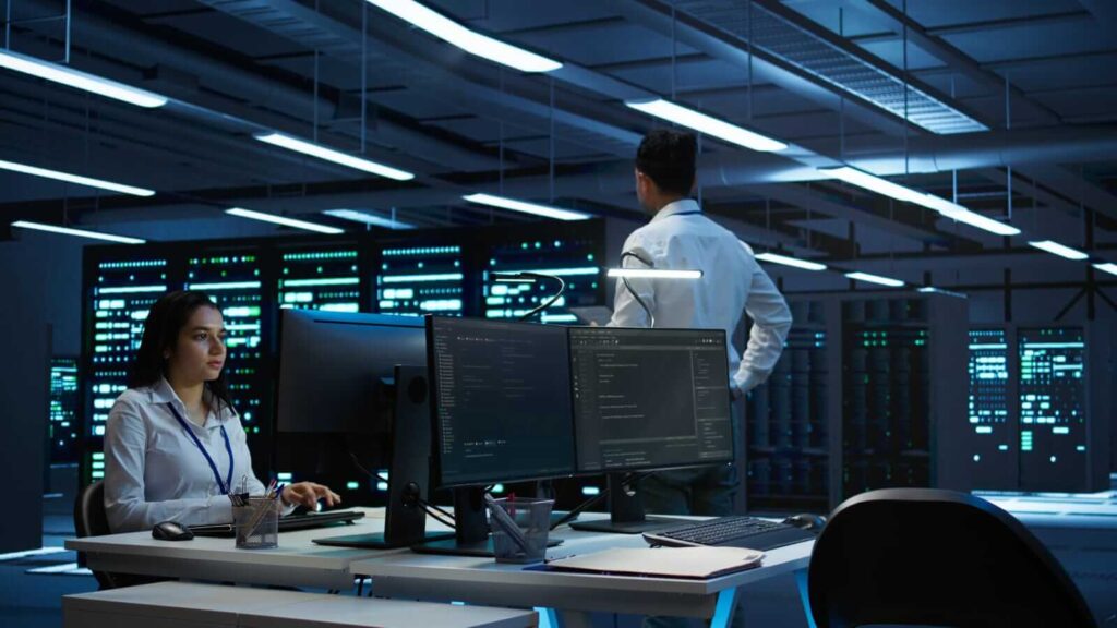 A woman works at a computer in a modern office with large screens. A man stands in the background, looking at servers.