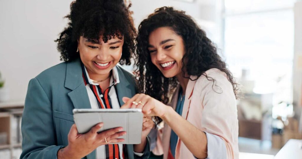Two women standing indoors, smiling and looking at a tablet device together—perhaps exploring new email hosting solutions.