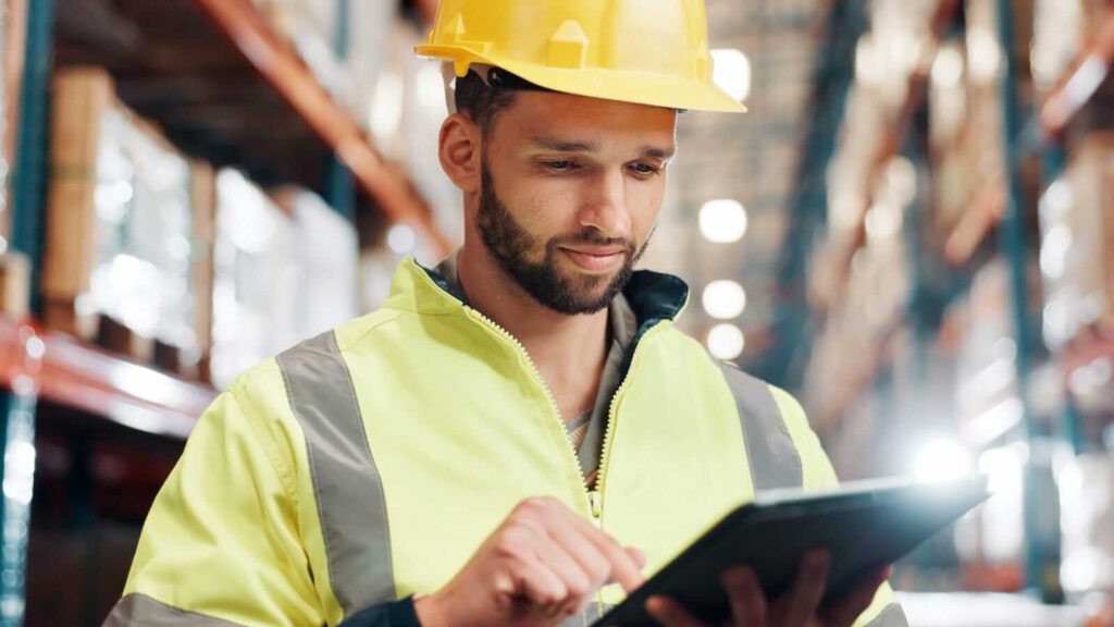 A man wearing a yellow hard hat and high-visibility jacket uses a tablet in a warehouse with shelves in the background, checking email hosting details for efficient communication.