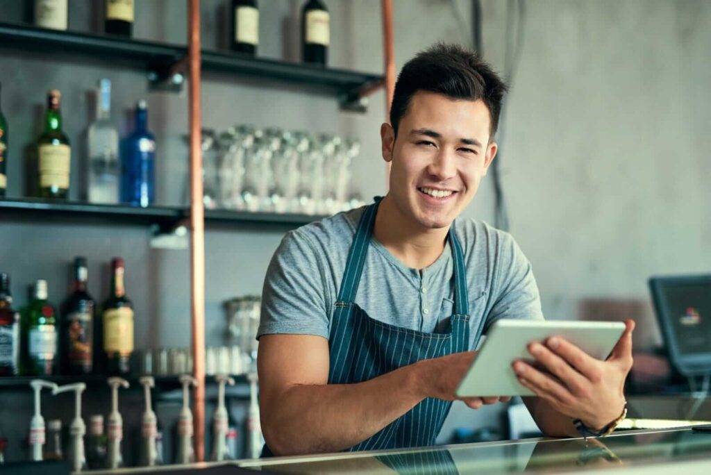 A young man in an apron stands behind a bar counter, smiling and holding a tablet, perhaps checking email hosting options, with bottles and glasses on shelves in the background.