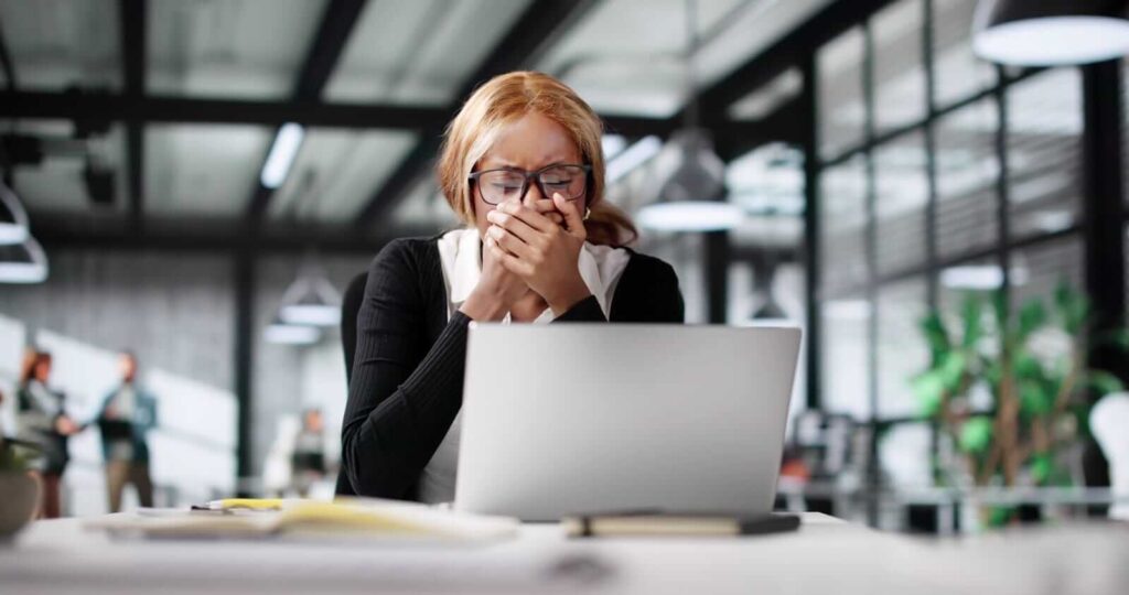 A woman wearing glasses sits at a desk with a laptop, covering her mouth with her hands and appearing distressed in a modern office setting.