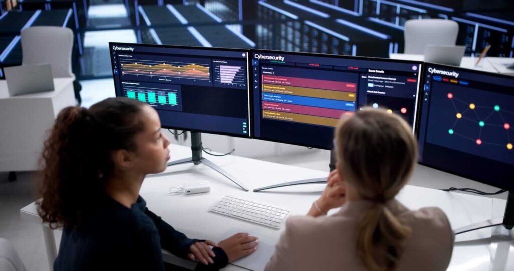 Two people sit at a desk analysing cybersecurity data on dual computer monitors displaying charts and threat alerts in an office setting.