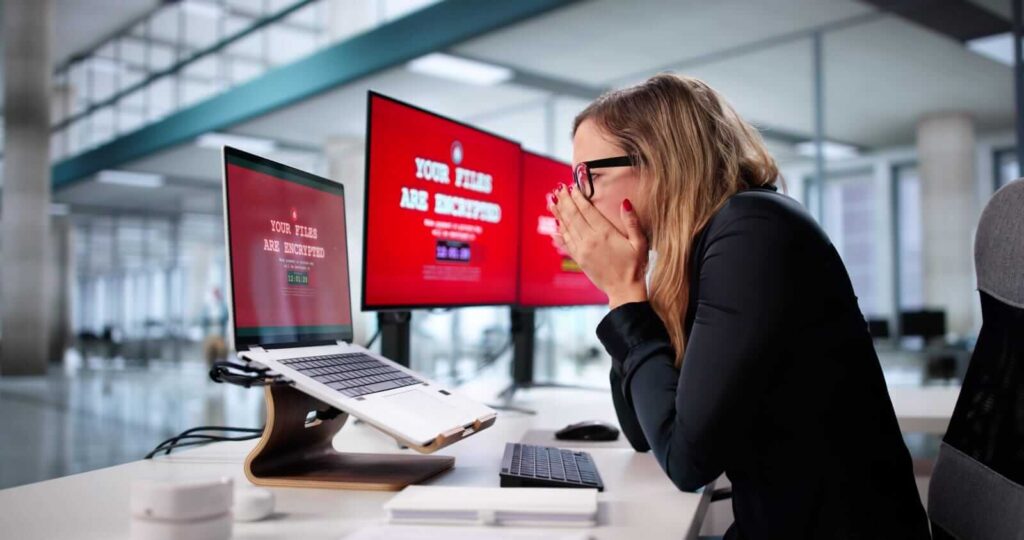 A woman in an office reacts with shock whilst looking at computer screens displaying a ransomware message stating "Your files are encrypted.